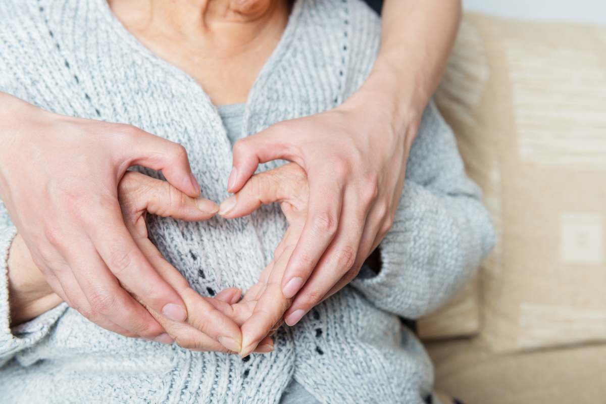 Donna anziana e persona dietro di lei che formano due cuori sovrapposti con la mano 