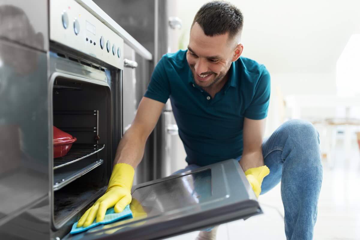 ragazzo con polo verde e guanti gialli che pulisce la porta di vetro del forno con pezza verde