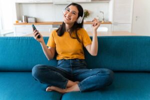 Ragazza felice con t-shrit gialla e jeans seduta sul divano con le cuffie e telecomando in mano